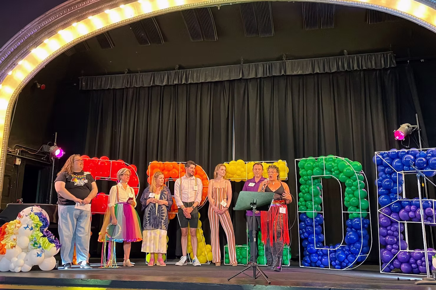 Speakers presenting on stage at the 2023 ILEA Seattle Pride Extravaganza in front of rainbow balloon letters
