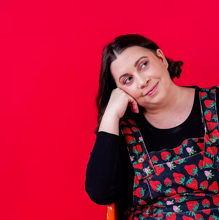 Ryan from Cute Story Events resting their head on their hand, smiling softly against a bright red background, wearing a black top with a strawberry-patterned dress