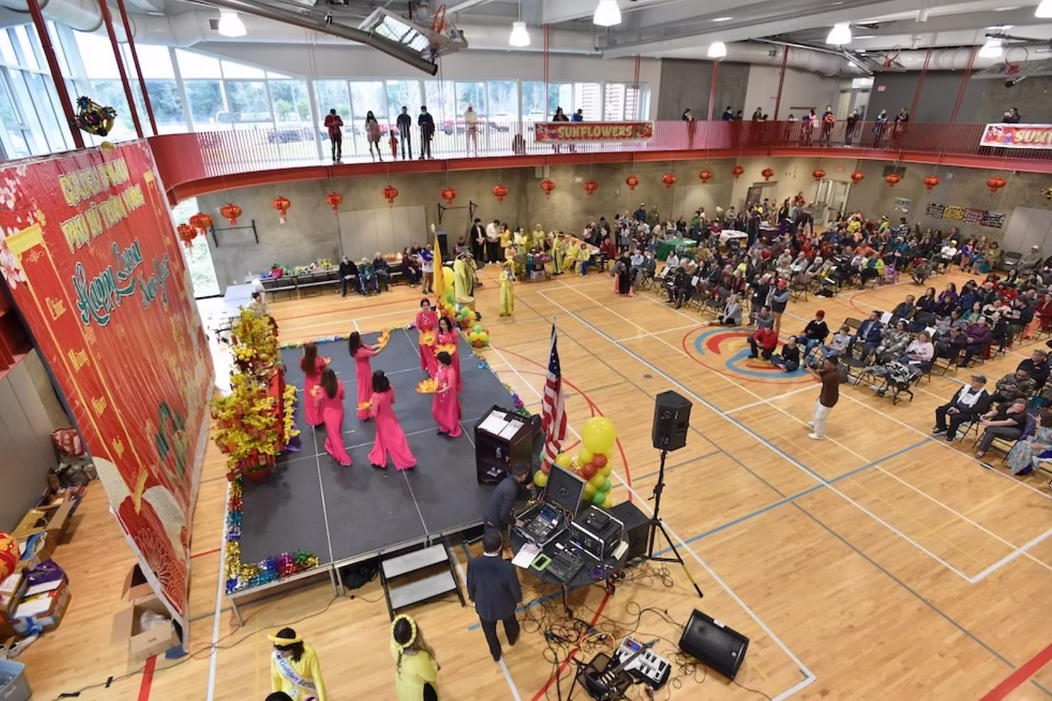 Wide view of a Vietnamese Tet Lunar New Year celebration featuring traditional performers on stage, red lantern decorations, and a large audience gathered inside a community center.