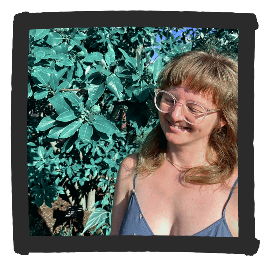 Caitlin Whyte smiling in front of leafy green plants, wearing glasses and a sundress