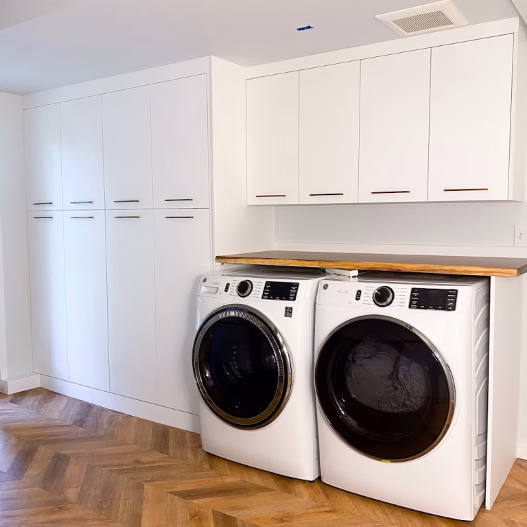 Modern laundry room with white frameless cabinets, built-in storage, and overhead lighting for a clean, organized look. Design by Iconic Cabinetry.