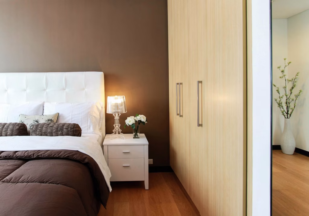Modern built-in bedroom closet with light wood panel doors beside a white upholstered bed and nightstand, styled with neutral tones and soft lighting