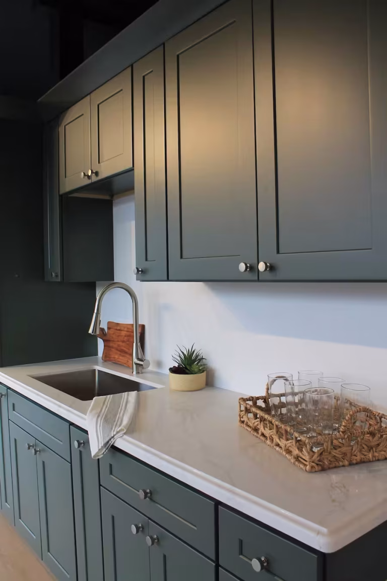 Modern green shaker-style kitchen cabinets with white countertop, stainless steel sink, striped dish towel, potted succulent, and a woven tray holding drinking glasses