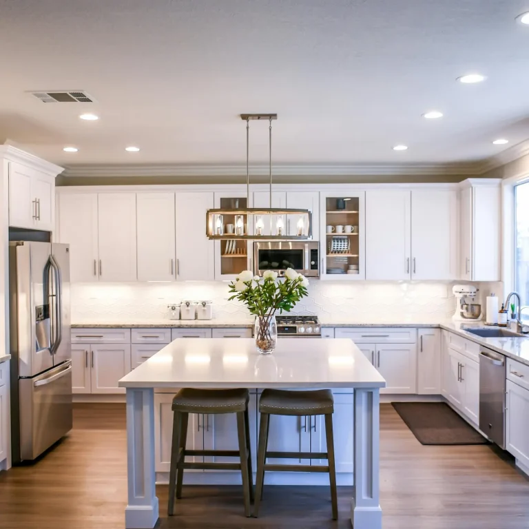 Bright modern kitchen with white shaker cabinets, stainless steel appliances, a center island with bar stools, and a decorative vase of flowers under a contemporary chandelier