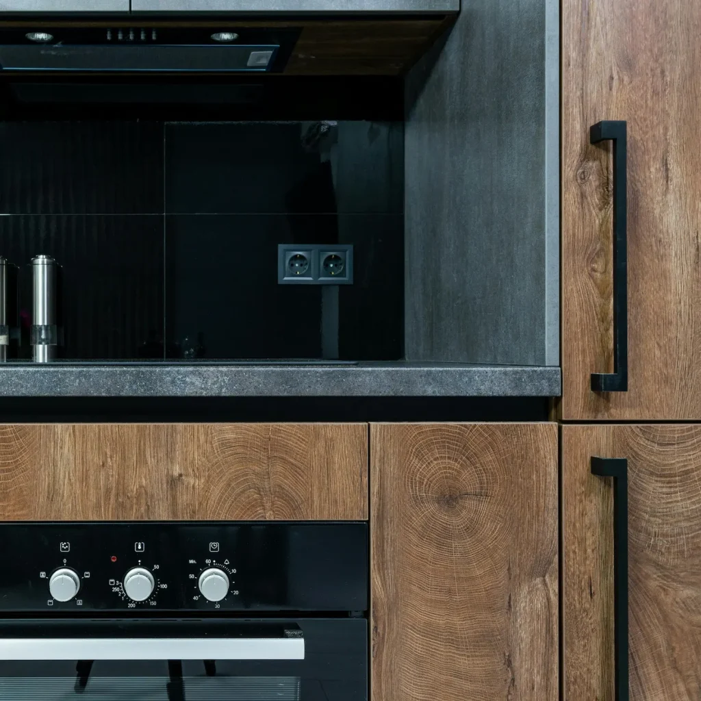 Modern kitchen with slim shaker-style wooden cabinets, black hardware, a built-in oven, and a black glass backsplash featuring a potted plant and salt and pepper shakers