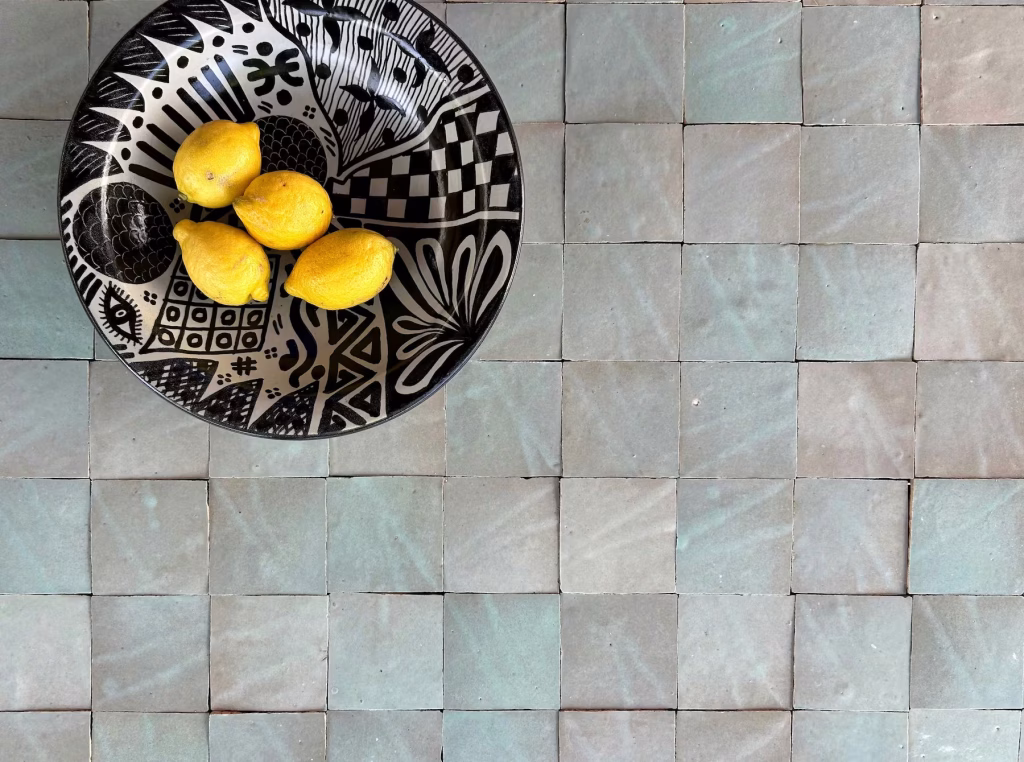 Top view of a patterned black and white bowl with four yellow lemons placed on a surface of square, muted green and gray handmade tiles.