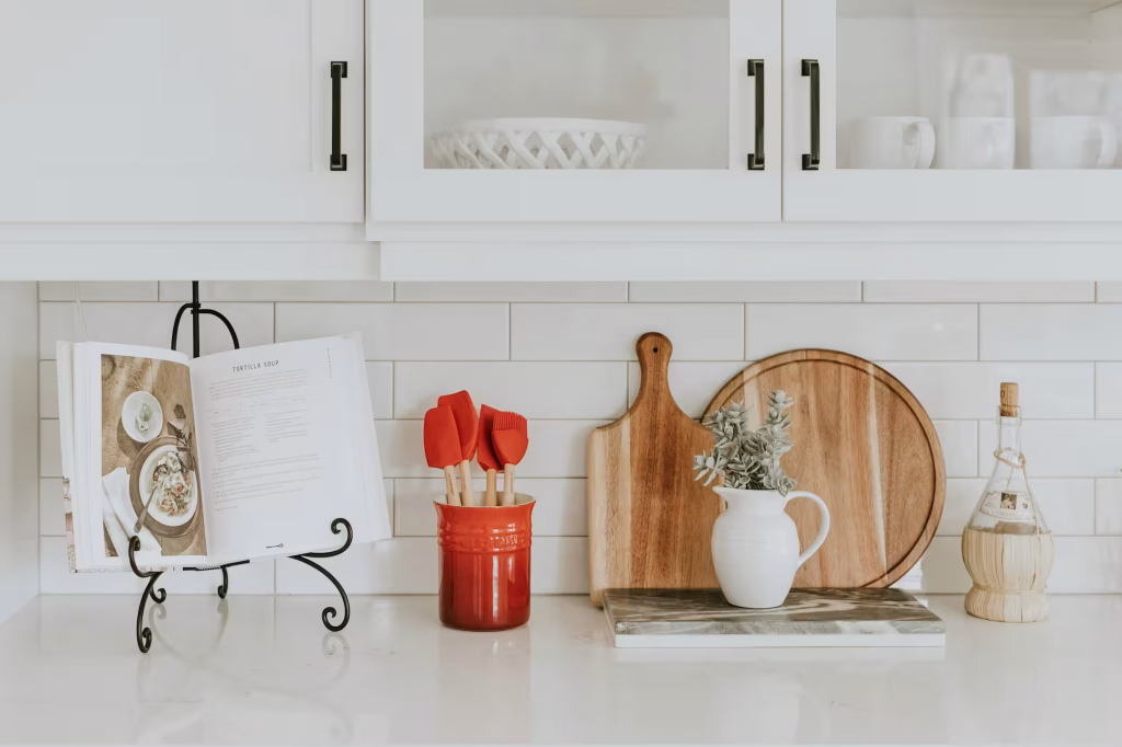 White shaker cabinets with subway tile backsplash, cookbook display, and red kitchen utensils on quartz countertop
