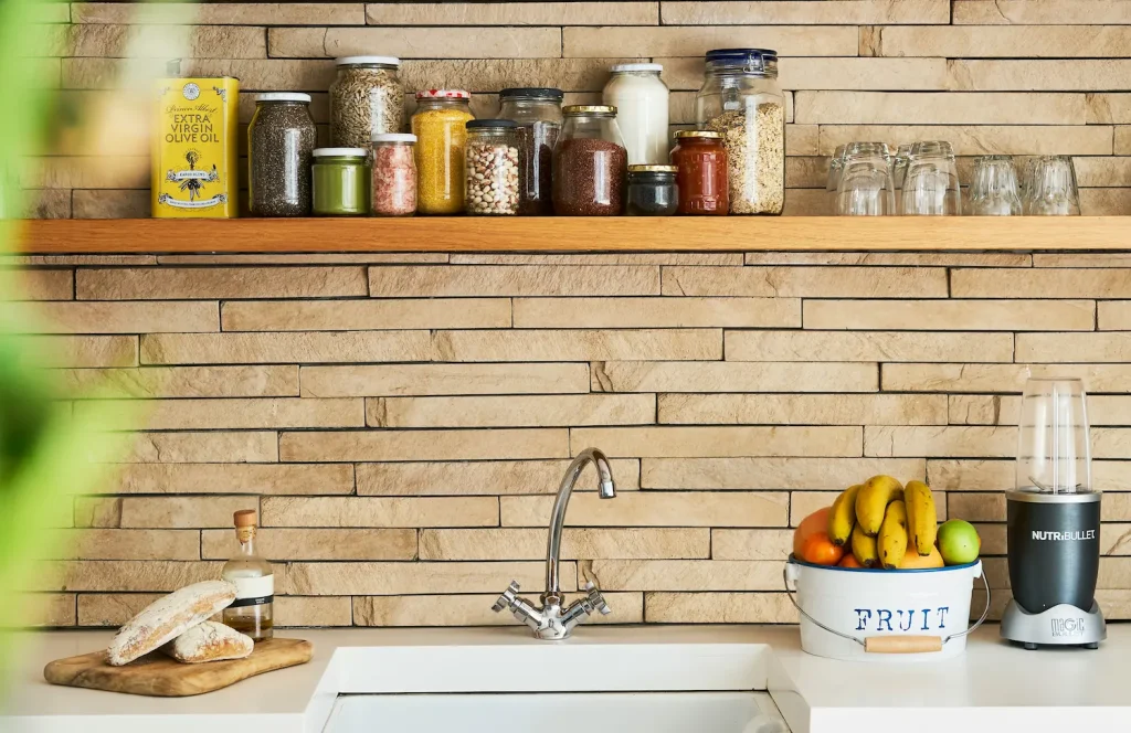 Rustic kitchen with exposed stone backsplash, wood shelf, and pantry jars in a natural-toned remodel design